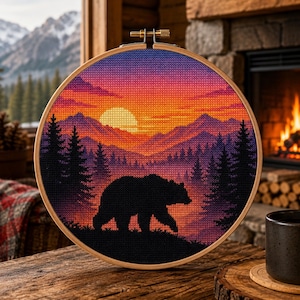 Puede incluir: Un bastidor de bordado circular de madera que contiene un patrón de punto de cruz de la silueta de un oso negro caminando por un bosque. El fondo presenta una puesta de sol vibrante con tonos naranja, amarillo y morado sobre montañas.