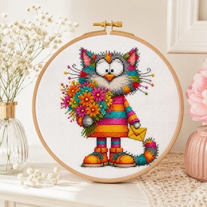 Peut inclure: Un tambour à broder circulaire en bois contenant un motif au point de croix représentant un chat gris avec des taches de fourrure multicolores. Le chat porte un haut rayé, tient un grand bouquet de fleurs vives et porte une enveloppe jaune.