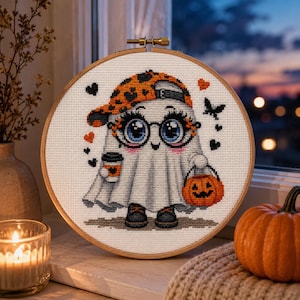 May include: A cross-stitch embroidery in a round wooden hoop featuring a white ghost character wearing an orange and black leopard print baseball cap, large round glasses, and black shoes. The ghost holds a coffee cup and a jack-o'-lantern bucket.