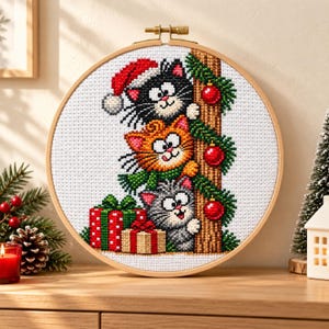 Op de afbeelding: Een ronde houten borduurring met een kruissteekpatroon van drie katten die verticaal op elkaar gestapeld tegen een boomstam staan. De bovenste kat draagt een rode kerstmuts. Groene guirlandes met rode kerstballen omwikkelen de boom, twee cadeaus onderaan.