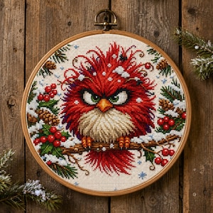 May include: A circular wooden embroidery hoop containing a cross-stitch pattern of a grumpy red bird with green eyes and a yellow beak. The bird is perched on a brown branch surrounded by green pine needles, brown pinecones, and red berries.
