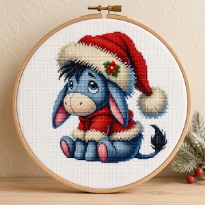 May include: A cross-stitch embroidery in a round wooden hoop featuring a blue donkey character wearing a red Santa hat with white trim and a small green and red flower detail, and a red sweater with white trim, set against a white fabric background.