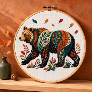 Op de afbeelding: Een ronde houten borduurring met een kruissteekpatroon van een bruine beer. De beer is versierd met kleurrijke blad- en bloemmotieven in tinten oranje, petrol en geel. Verspreide blaadjes zweven boven de beer op de witte stof.
