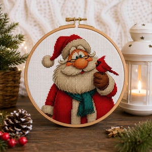 May include: A circular wooden embroidery hoop containing a cross-stitch pattern of Santa Claus wearing a red hat, a red coat, and a teal scarf. Santa has a large white beard, glasses, and is holding a small red cardinal bird on one hand.