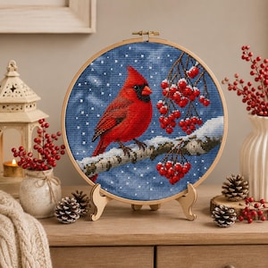 Può includere: Ricamo a punto croce di un cardinale rosso appollaiato su un ramo innevato con bacche rosse. L'opera è esposta in un cerchio di legno e posta su un supporto di legno. Lo sfondo è un cielo blu con neve.