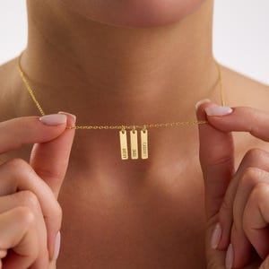 May include: A gold-colored necklace with three rectangular pendants. Each pendant is engraved with a name: Avery, Jade, and Cadence. The necklace is held by a person.