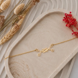 May include: A gold-toned necklace featuring a central pendant composed of two parallel, angled bars connected to four small circular loops. The delicate chain extends from both sides of the geometric centerpiece, which has a polished, reflective finish.