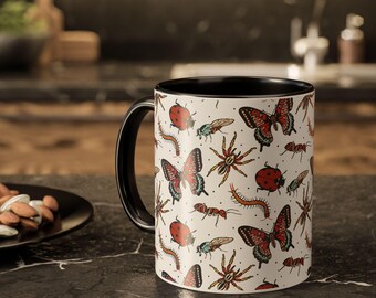 Puede incluir: Taza de cerámica blanca con interior y asa negros. La taza está cubierta con un patrón repetido de mariposas rojas y negras, mariquitas, arañas, hormigas, ciempiés y moscas. La taza está sobre una encimera negra.