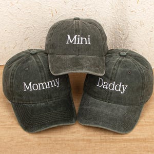Op de afbeelding: Drie donkergroene baseballpetten met wit geborduurde tekst. Eén pet zegt "Mini", een andere zegt "Mommy" en de derde zegt "Daddy". De petten zijn gerangschikt op een lichtbruin houten oppervlak.
