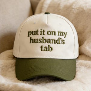 Puede incluir: Gorra de béisbol con corona blanca y visera verde oliva. La frase "put it on my husband's tab" está bordada en hilo verde oliva en la parte delantera. El gorro está sobre una suave manta color crema.