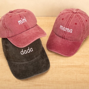 Puede incluir: Se muestran tres gorras de béisbol en tonos rojos y negros. Las gorras rojas tienen las palabras "mini" y "mama" bordadas en blanco. La gorra negra tiene la palabra "dada" bordada en blanco. Las gorras están sobre una superficie de madera.