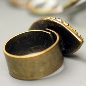 May include: A brass ring with a large, oval-shaped stone set in the top. The stone is surrounded by small, clear rhinestones.