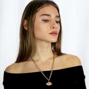 May include: A gold chain necklace with an evil eye pendant. The pendant is made of gold and has a blue stone in the centre. The necklace is worn by a person with long brown hair.