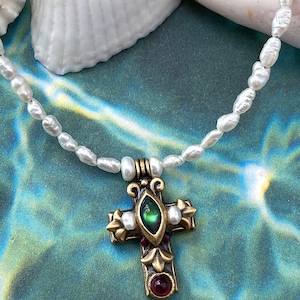 Puede incluir: Un collar de perlas blancas con un colgante de cruz dorada. La cruz tiene una piedra verde e iridiscente en el centro y una piedra roja en la parte inferior.