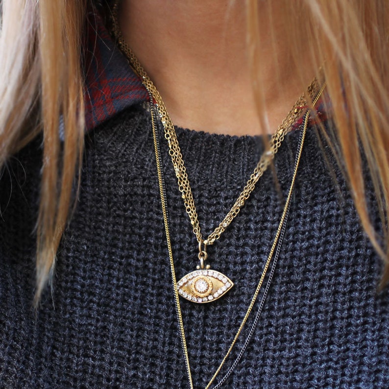 May include: A gold chain necklace with a large eye pendant featuring a clear crystal in the centre. The necklace is layered over a black sweater.