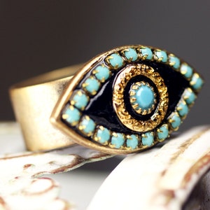 May include: A gold ring with a blue evil eye design. The eye is surrounded by a gold frame and turquoise stones.