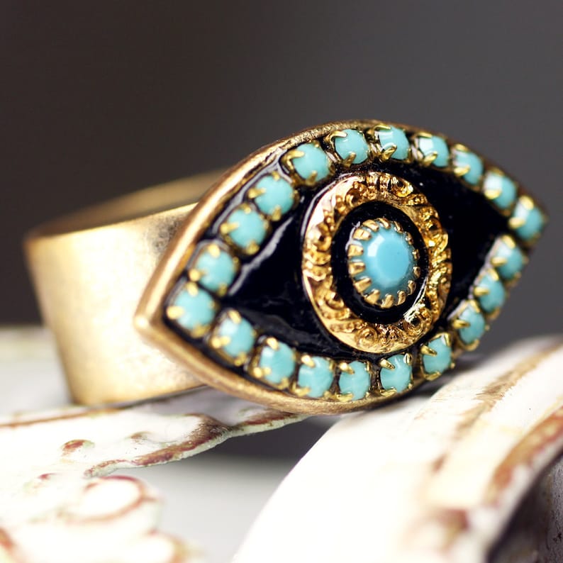 May include: A gold ring with a blue evil eye design. The eye is surrounded by a gold frame and turquoise stones.