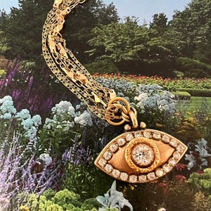 May include: A gold chain necklace featuring a large, eye-shaped pendant. The pendant is set with a large, clear diamond in the centre and smaller diamonds around the edge. The pendant is surrounded by a gold frame.