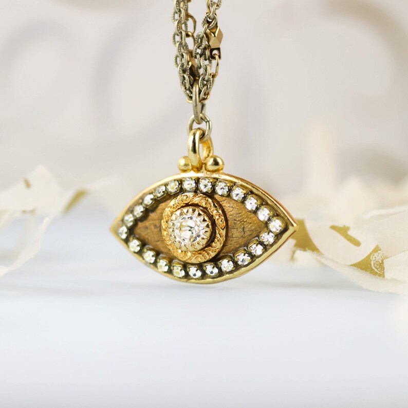 May include: Gold eye pendant necklace with a clear gemstone in the centre and a wood-like background. The pendant is surrounded by a circle of small clear gemstones.