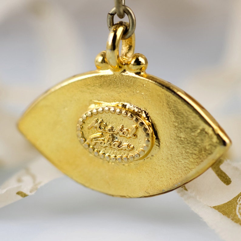 May include: Gold-tone eye charm pendant featuring a Michael Follain logo. The charm is shaped like an eye with a gold-tone border and a blue centre.