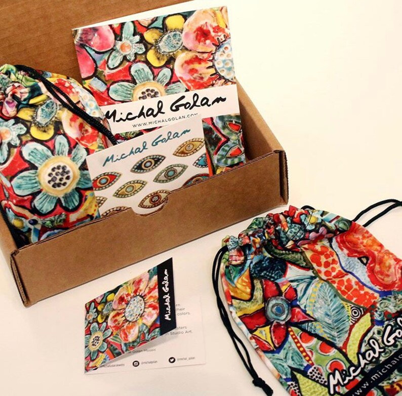 May include: A brown cardboard box with a colorful patterned fabric bag inside. The bag has a drawstring closure and is decorated with a floral design. The box also contains a small card with the text "Michal Golan" and a website address.