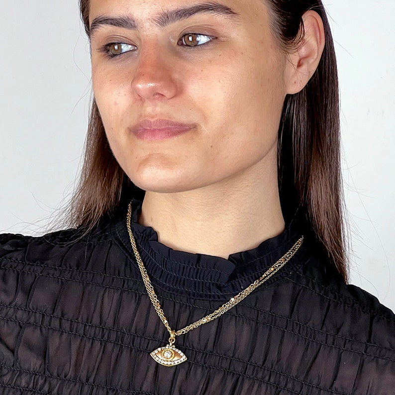 May include: A gold chain necklace with a small, eye-shaped pendant. The pendant is decorated with small, clear stones.