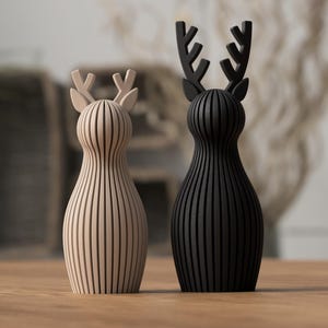 Puede incluir: Dos figuras decorativas de ciervos. Una es de color beige claro, la otra es negra. Ambas tienen un dise&ntilde;o de rayas verticales y astas estilizadas. Se colocan sobre una superficie de madera, con un fondo borroso.