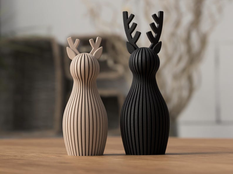 Puede incluir: Dos figuras decorativas de ciervos. Una es de color beige claro, la otra es negra. Ambas tienen un dise&ntilde;o de rayas verticales y astas estilizadas. Se colocan sobre una superficie de madera, con un fondo borroso.
