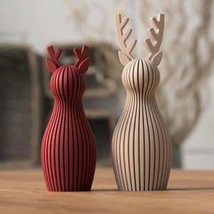 Puede incluir: Dos figuras de renos de madera. Una es de color rojo intenso y la otra de tono madera natural. Ambas tienen un dise&ntilde;o de rayas verticales y astas. Se exhiben sobre una superficie de madera.