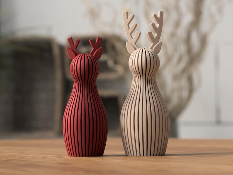Puede incluir: Dos figuras de renos de madera. Una es de color rojo intenso y la otra de tono madera natural. Ambas tienen un dise&ntilde;o de rayas verticales y astas. Se exhiben sobre una superficie de madera.