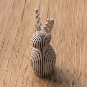 Puede incluir: Figura de reno de madera marr&oacute;n claro con un dise&ntilde;o de rayas verticales. El reno tiene un cuerpo redondeado y astas detalladas. La figura est&aacute; sobre un fondo de madera.