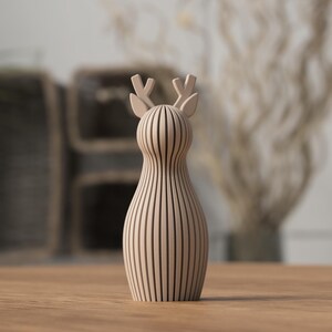 Puede incluir: Figura decorativa de ciervo de madera en color marr&oacute;n claro. El ciervo tiene un cuerpo esbelto y peque&ntilde;as astas. La figura est&aacute; hecha de listones de madera verticales, lo que le da un aspecto rayado. Se encuentra sobre una superficie de madera.