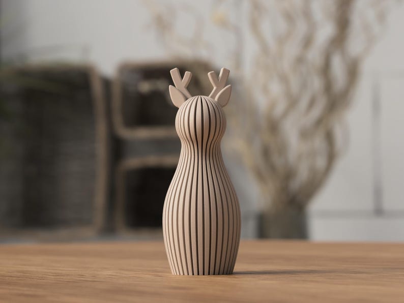 Puede incluir: Figura decorativa de ciervo de madera en color marr&oacute;n claro. El ciervo tiene un cuerpo esbelto y peque&ntilde;as astas. La figura est&aacute; hecha de listones de madera verticales, lo que le da un aspecto rayado. Se encuentra sobre una superficie de madera.