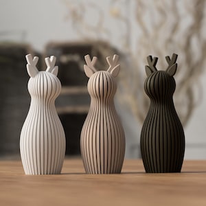 Puede incluir: Tres figuras decorativas de ciervos en blanco, beige y marr&oacute;n oscuro. Cada ciervo tiene un cuerpo a rayas verticales y astas. Las figuras se exhiben sobre una superficie de madera.