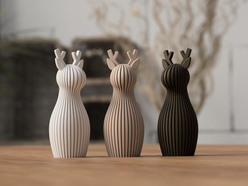 Puede incluir: Tres figuras decorativas de ciervos en blanco, beige y marr&oacute;n oscuro. Cada ciervo tiene un cuerpo a rayas verticales y astas. Las figuras se exhiben sobre una superficie de madera.
