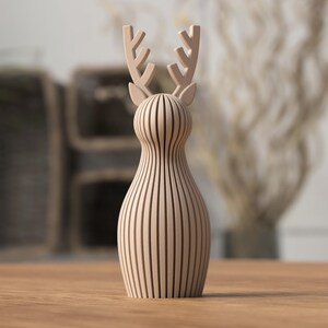 Puede incluir: Una figura de ciervo de madera de color marr&oacute;n claro. El ciervo tiene un cuerpo delgado con l&iacute;neas verticales y un par de cuernos. La figura est&aacute; sobre una superficie de madera.