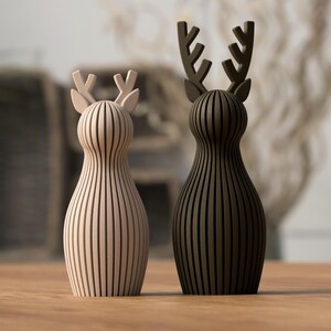 Puede incluir: Dos figuras de ciervo de madera, una beige claro y otra marr&oacute;n oscuro, de pie una al lado de la otra. Cada una tiene un cuerpo a rayas verticales y astas estilizadas. Las figuras est&aacute;n sobre una superficie de madera, con un fondo borroso.