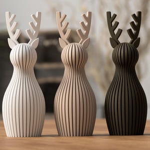 Puede incluir: Tres figuras decorativas de madera de renos en tonos blanco, beige y marr&oacute;n oscuro. Cada una tiene un cuerpo con rayas verticales y astas estilizadas. Las figuras se exhiben sobre una superficie de madera.