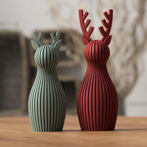 Puede incluir: Dos figuras decorativas de renos. Una es verde salvia, la otra es roja. Ambas tienen un dise&ntilde;o de rayas verticales y astas. Se colocan sobre una superficie de madera, con un fondo borroso.