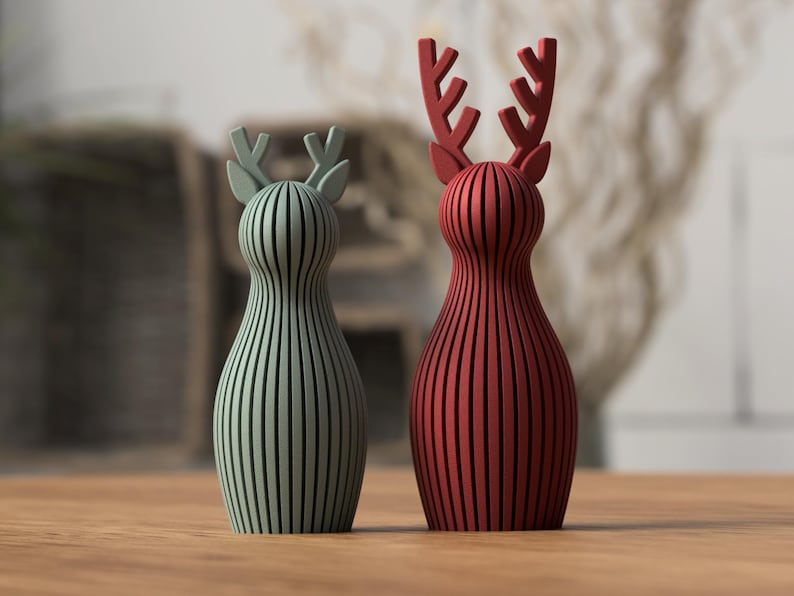 Puede incluir: Dos figuras decorativas de renos. Una es verde salvia, la otra es roja. Ambas tienen un dise&ntilde;o de rayas verticales y astas. Se colocan sobre una superficie de madera, con un fondo borroso.