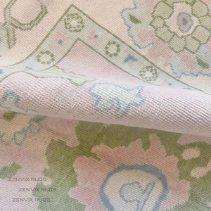 May include: A folded area rug featuring a soft pink background with intricate floral patterns in sage green and light blue. The textile displays a repeating geometric border and organic botanical motifs. The text ZENVIX RUGS is visible.