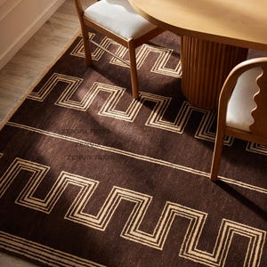 May include: A rectangular area rug featuring a deep brown base color with a repeating cream-colored geometric Greek key pattern. The rug displays the text ZENVIK RUGS printed in a subtle font. The design includes horizontal lines and borders.
