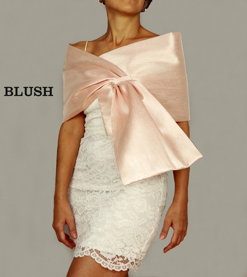 A blush pink satin shawl with a large bow tied in the front. The shawl is draped over a white lace dress.