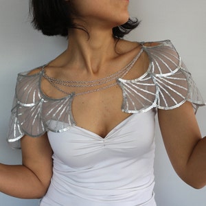 Op de afbeelding: Een zilveren schouderstuk met pailletten en een delicate kettingketting. Het schouderstuk is gemaakt van een transparante stof met een gegolfde rand.