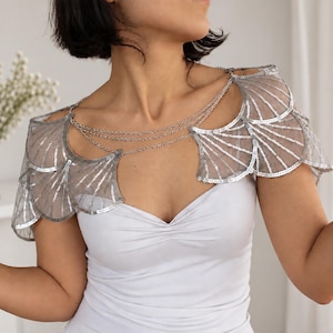 Model wearing a silver sequin bridal capelet over a white strapless dress, showing the wing-shaped embroidered tulle shoulders and delicate aluminum chain drapes across the neckline.
