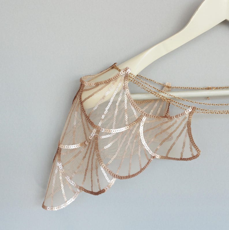 Close-up of the rose-gold sequin embroidered bridal shoulder capelet on a hanger, highlighting the geometric embroidery, sheer tulle and chain details.