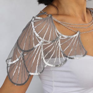 Close-up of a silver sequin bridal shoulder capelet, showing layered wing-shaped panels, geometric embroidery on sheer gray tulle, and delicate aluminum chains draped across the neckline.