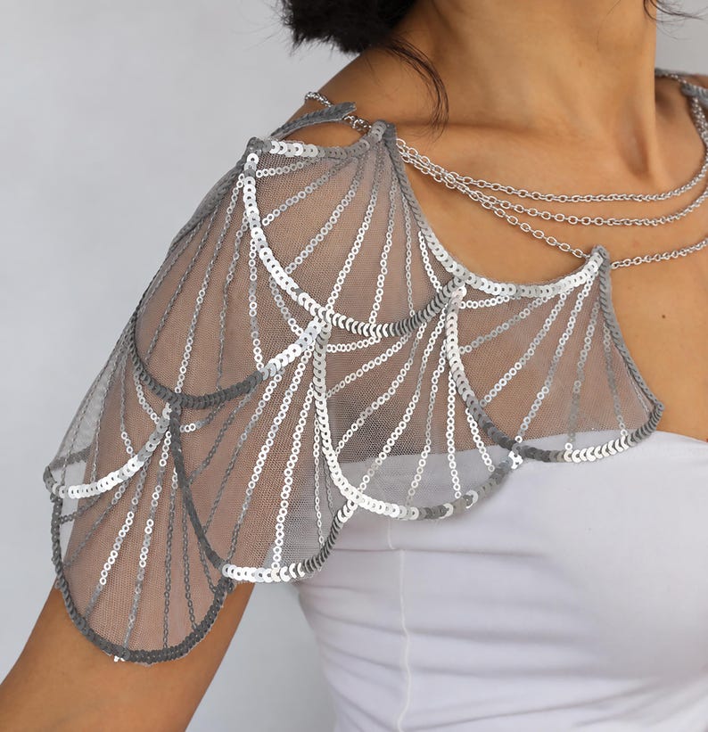 Close-up of a silver sequin bridal shoulder capelet, showing layered wing-shaped panels, geometric embroidery on sheer gray tulle, and delicate aluminum chains draped across the neckline.