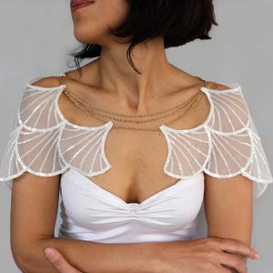 Model wearing the silver sequin bridal capelet over a white strapless dress, showing the shoulder wings and decorative chains from a three-quarter angle.
