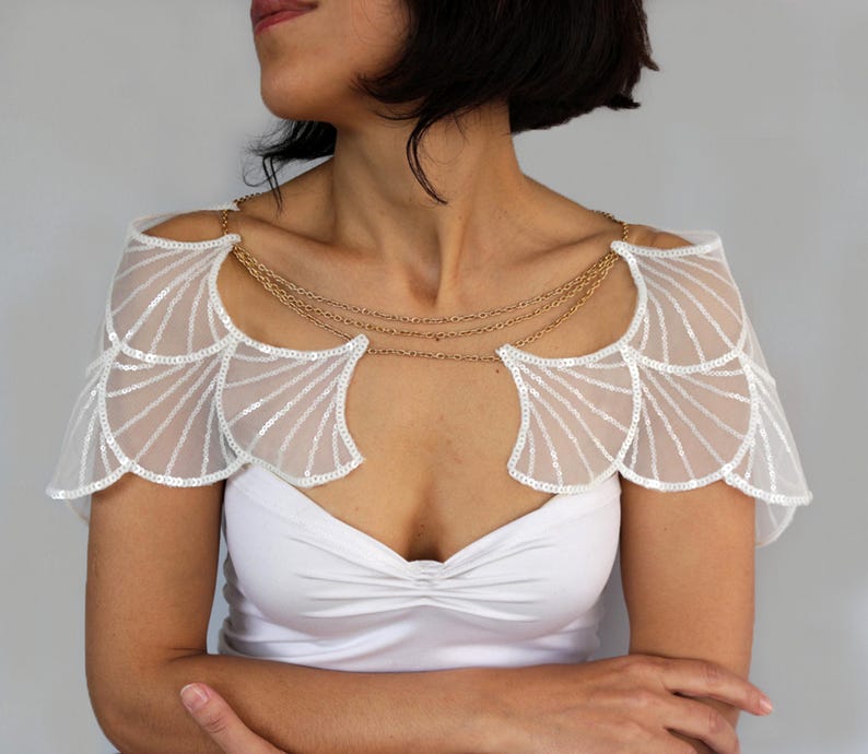 Model wearing the silver sequin bridal capelet over a white strapless dress, showing the shoulder wings and decorative chains from a three-quarter angle.